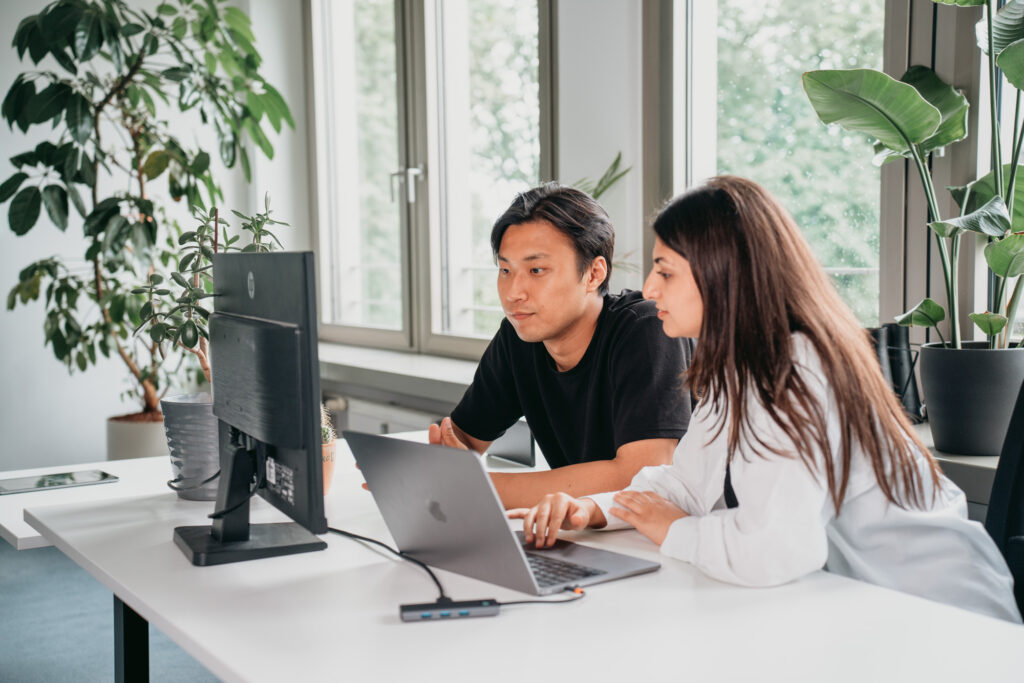 Zwei Personen arbeiten in einer Büroumgebung an einem Schreibtisch mit Computern zusammen. Die Person links im schwarzen Hemd schaut auf den Monitor, während die Person rechts im weißen Hemd an einem Laptop arbeitet. Topfpflanzen und große Fenster sorgen für einen hellen Hintergrund, während sie sich auf die Power App Entwicklung konzentrieren.