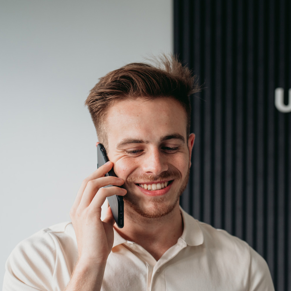 Ein junger Mann mit kurzen braunen Haaren und leichtem Bart lächelt und telefoniert mit einem Smartphone. Er trägt ein helles Hemd mit Kragen und steht vor einem neutralen Hintergrund mit einem vertikalen schwarzen Panel auf der rechten Seite. Vielleicht bespricht er gerade sein neuestes Projekt in Power App Entwicklung.