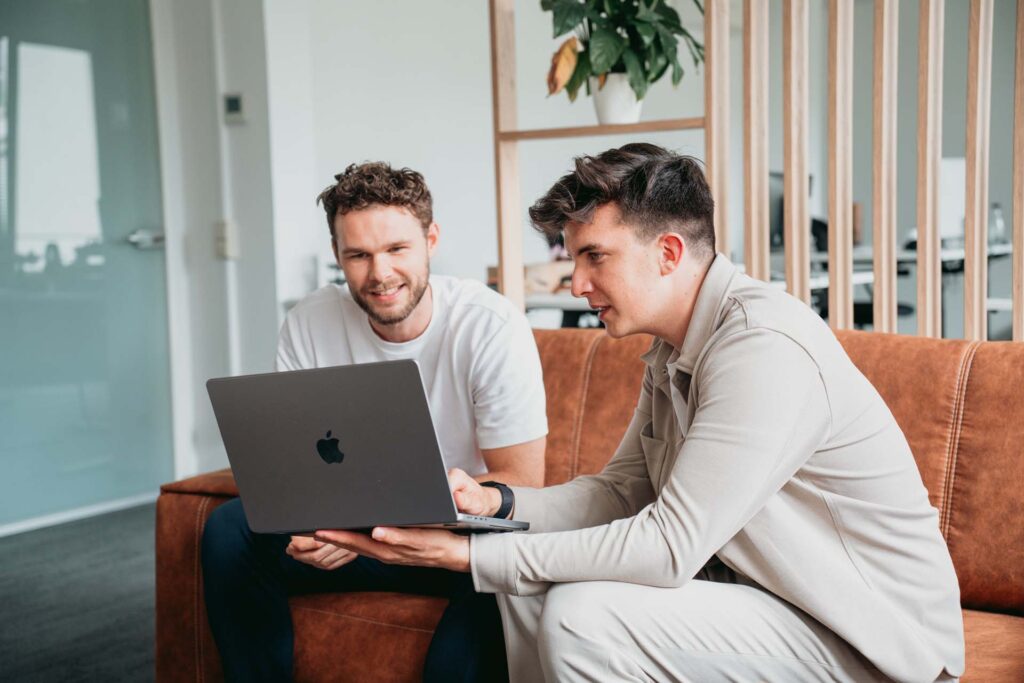 Zwei Männer sitzen auf einem braunen Sofa und schauen auf einen Laptop-Bildschirm. Einer der Männer hält den Laptop, während beide lächeln und sich mit dem Inhalt auf dem Bildschirm beschäftigen. Wahrscheinlich erkunden sie Microsoft Power Apps. Im Hintergrund steht eine Pflanze und durch eine Glaswand ist eine Büroumgebung sichtbar.