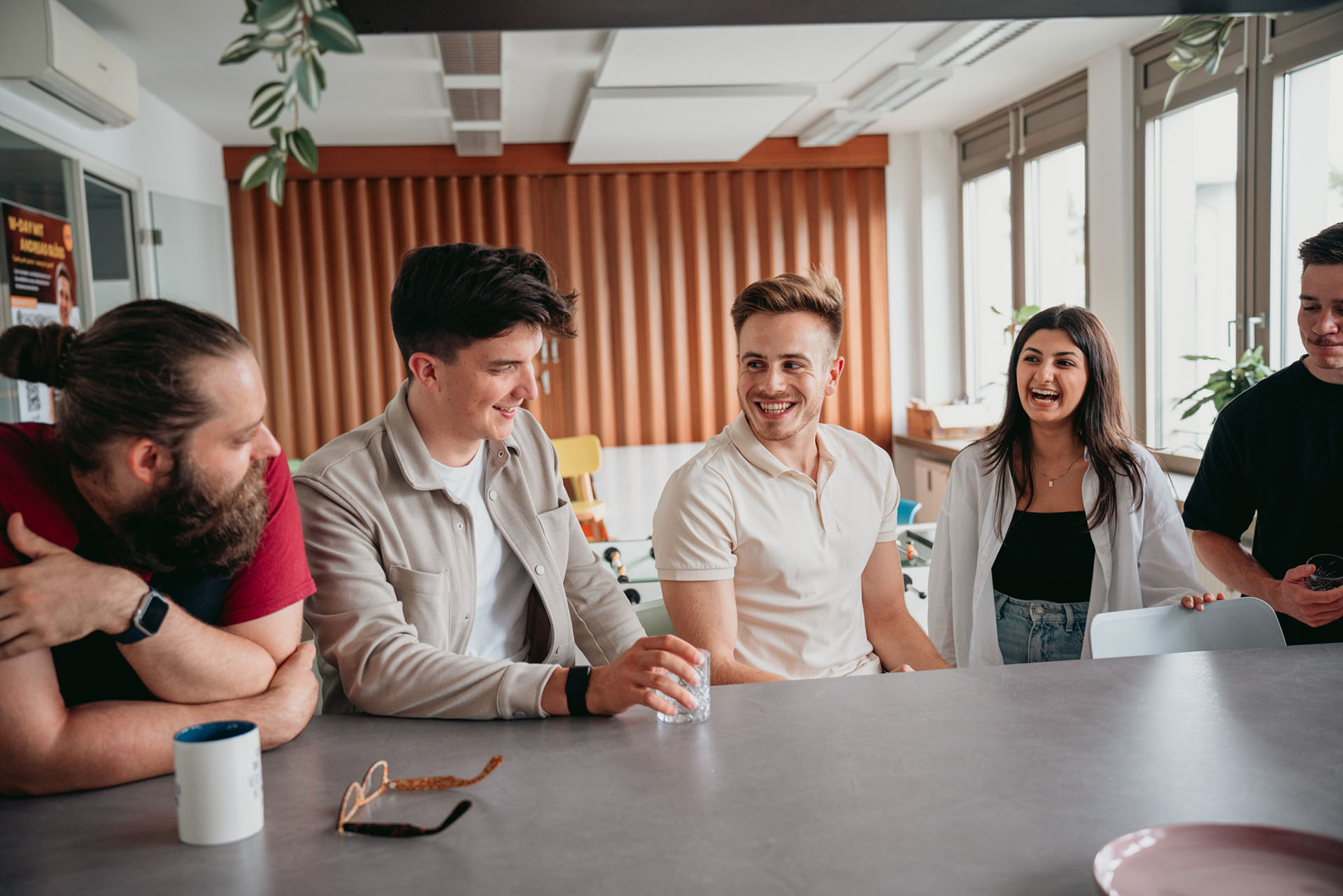 Eine Gruppe von fünf Personen sitzt in einem modernen Raum um einen Tisch herum und lacht und unterhält sich. Eine Person lehnt am Tisch, auf dem Gegenstände wie eine Kaffeetasse, Gläser und Notizen zur Microsoft Power Plattform verstreut liegen. Der Raum ist hell und das Tageslicht fällt durch die großen Fenster.